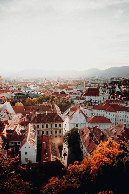 Panorama von Graz im Herbst – Blick über die Altstadt mit roten Dächern, historischen Gebäuden und herbstlich gefärbten Bäumen, im Hintergrund sanfte Hügel.